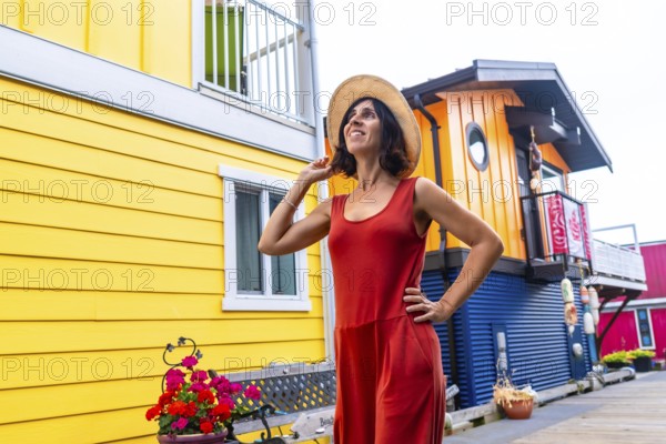 Happy female tourist wearing a straw hat and red dress is posing and smiling in front of the colorful floating houses of fisherman's wharf park in victoria, british columbia