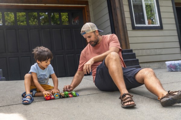 Father and son are sitting on the ground in their driveway, playing with a collection of toy cars, enjoying quality time together on a sunny day