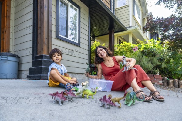 Mother and son sitting on the ground in their front yard, enjoying a sunny summer day while playing with a colorful collection of dinosaur toys, fostering imagination and joy
