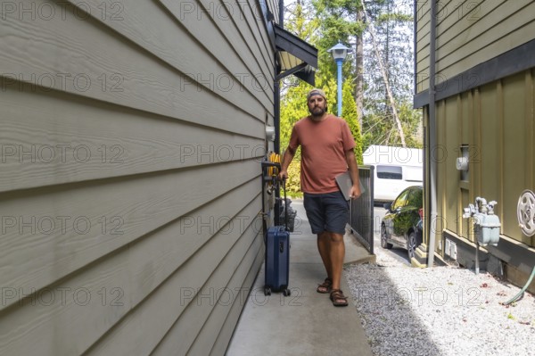 Casually dressed man walking outside his house, pulling a suitcase while holding a laptop, eagerly starting his summer vacation and ready for an exciting getaway