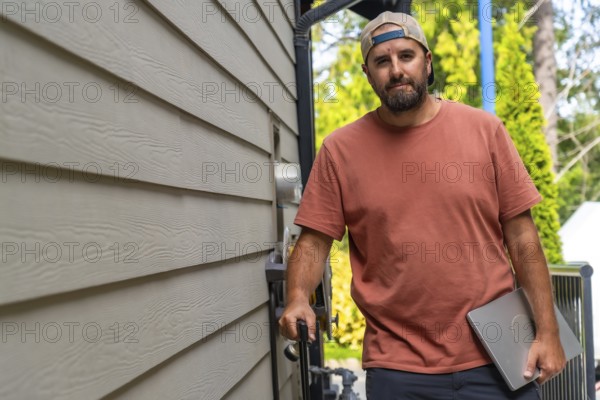 Maintenance worker walking alongside a residential building, carrying a laptop and various tools, dressed in casual attire with a cap, ready for service and repair tasks