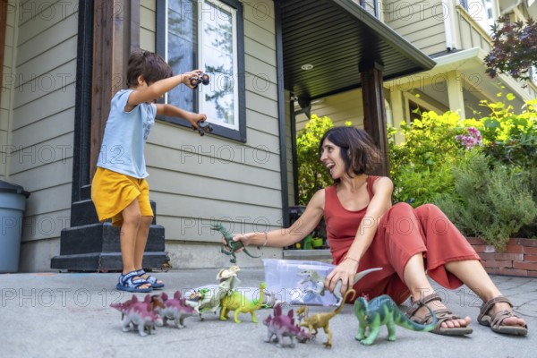 Happy mother and son enjoying quality time together, playing with dinosaur toys outside their house on a sunny day, fostering imagination and family bonding