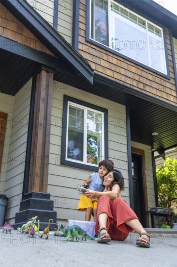 Mother and her son are sitting on the driveway of their house, playing with dinosaur toys on a sunny summer day, creating a heartwarming scene of family bonding