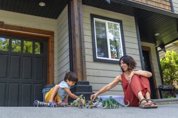 Mother and son sitting on the pavement in front of their house, joyfully playing with colorful dinosaur toys on a bright, sunny summer day, enjoying precious moments of childhood together