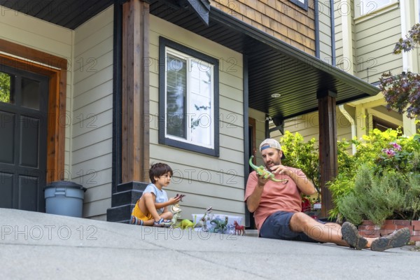 Father and son enjoying quality time together, bonding over dinosaur toys while sitting on their driveway, creating joyful memories outside their modern home in a suburban neighborhood