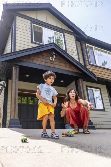 Mother and son are crouching down, playing with toy cars on the driveway in front of their house, enjoying quality time together on a sunny summer day