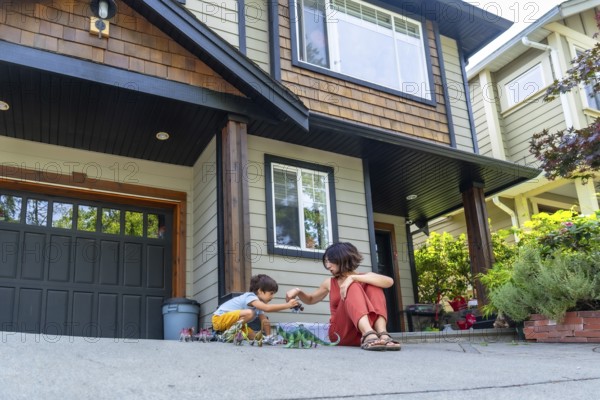 Mother and son bonding over dinosaur toys, enjoying quality time together on the driveway of their suburban home, fostering imagination and family connection in a playful setting