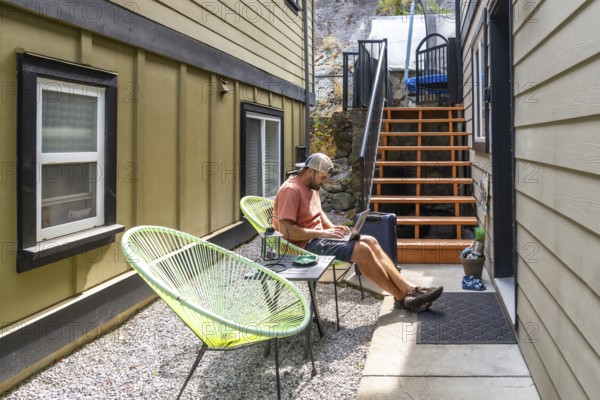 Young man working remotely on a laptop, lounging comfortably in a patio chair in his backyard, soaking up the warm summer sun and enjoying the relaxed outdoor atmosphere