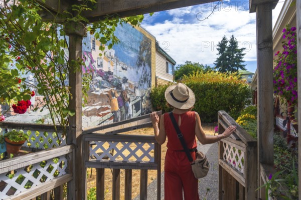 Female tourist in a straw hat and red jumpsuit opening a wooden gate to admire a vibrant mural depicting the city's history in chemainus, vancouver island, on a sunny summer day
