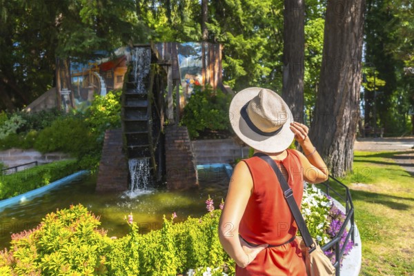 Tourist wearing a straw hat holds the brim while admiring a water wheel in a garden setting in chemainus, british columbia, on vancouver island, canada, during a sunny summer day