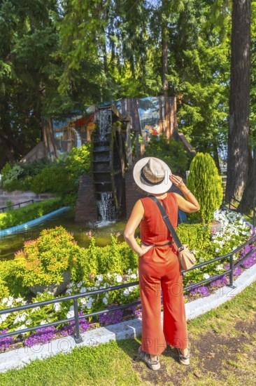 Female tourist wearing a straw hat and a jumpsuit is admiring a watermill and a colorful mural in a park in chemainus, british columbia, during summer