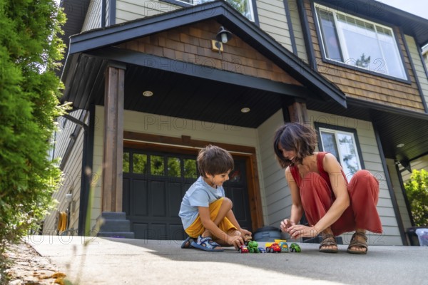 Mother and her young son are happily playing with toy cars on the driveway of their suburban home, enjoying quality time together on a sunny day