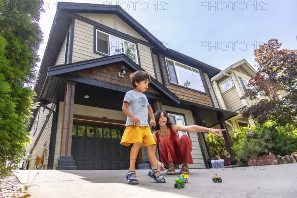 Mother crouches next to her son, pointing at toy cars on the driveway of their two story home, creating a heartwarming scene of family bonding and childhood play