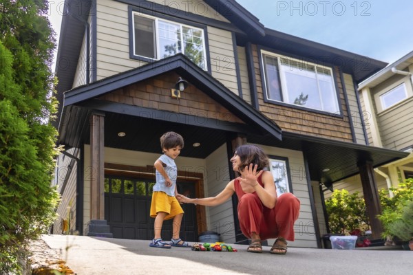 Mother interacts with her young son, playing with toy cars on the driveway of their modern suburban home, creating a heartwarming scene of family life and childhood joy