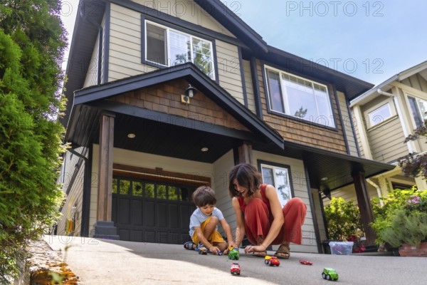 Mother and her young son are happily playing with toy cars on the driveway of their suburban home, enjoying quality time together on a sunny summer day