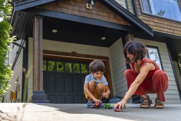 Mother and her young son are happily playing with toy cars on the driveway of their modern suburban home, enjoying quality time together on a sunny day