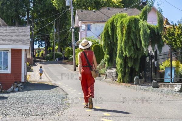 Tourist wearing a straw hat and orange jumpsuit walking along a yellow dotted line on a sunny summer day in chemainus, vancouver island, with houses and trees in the background