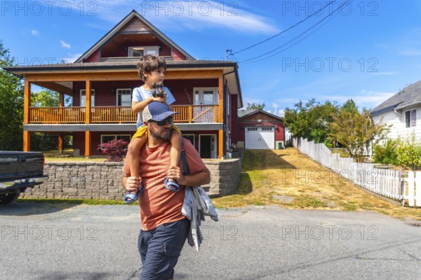 Father carrying son on shoulders while walking past a two story house with a porch and detached garage in a sunny suburban neighborhood in chemainus, british columbia