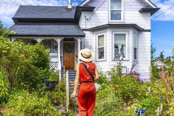 Tourist wearing a straw hat and an orange jumpsuit walks up a garden path towards a charming house in chemainus, british columbia, canada, on a sunny summer day