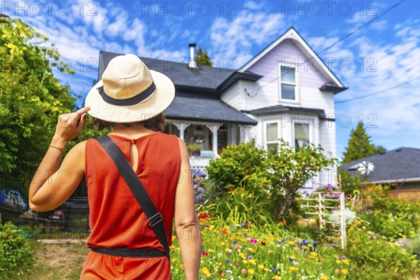 Female tourist wearing a straw hat, holding it with one hand, admiring a beautiful blooming garden and a charming house in chemainus, british columbia, canada, during a sunny summer day