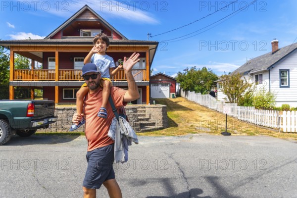 Father carrying his son on his shoulders waving in front of their house in chemainus, vancouver island, british columbia, canada, during a sunny summer day