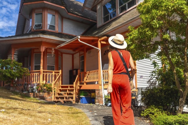 Tourist wearing a straw hat and a red jumpsuit is walking on a path near a traditional wooden house with an orange porch in chemainus, vancouver island, during a sunny summer day