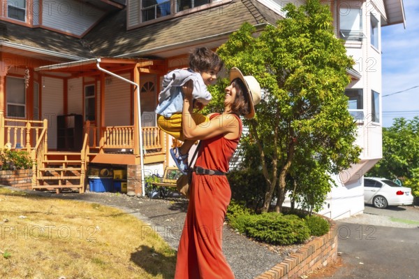 Happy mother wearing a straw hat and an orange jumpsuit lifting up her cheerful son in the yard of their house in chemainus, british columbia, canada