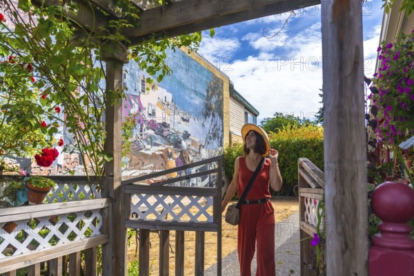 Female tourist wearing jumpsuit and straw hat walking through wooden gate and admiring colorful street art mural in chemainus, british columbia, canada, during sunny summer day