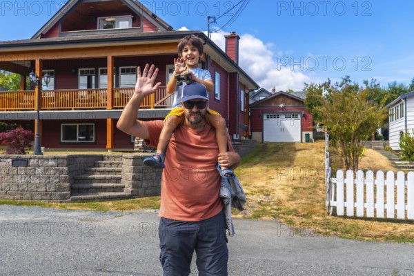 Father carrying his son on his shoulders, both waving, standing in front of their house in chemainus, vancouver island, british columbia, canada, enjoying a summer day