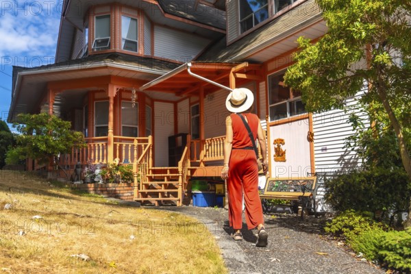 Tourist wearing a straw hat and orange jumpsuit walking along a gravel path beside a vibrant wooden house with an orange porch in chemainus, british columbia, on a sunny summer day