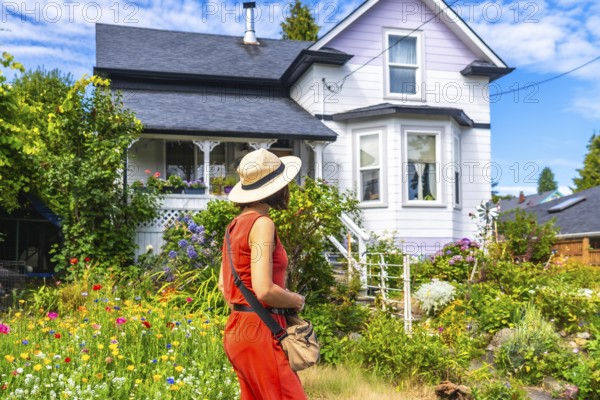Tourist wearing a straw hat and orange dress walking through a colorful blooming garden in front of a victorian house on a sunny summer day in chemainus, canada