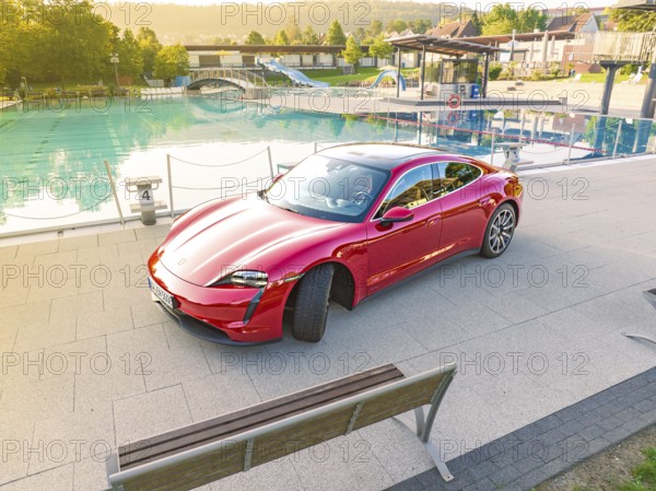 Red sports car next to a swimming pool and park bench in daylight, Porsche Taycan, Deer Carsharing, Calw, Germany