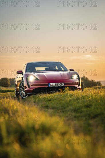 Red car is parked in a field at sunset, the sky glows orange in the background, Porsche Taycan, deer car sharing. Calw, Germany