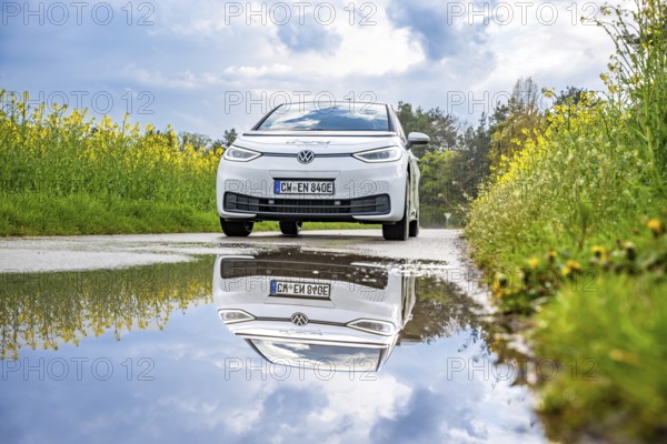 A white electric car on a country road, reflected in a puddle, surrounded by yellow flowers and blooming nature, ID3 VW, deer car sharing. Calw, Germany