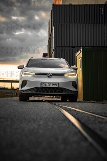 White car drives past container warehouse at sunset, dramatic urban scene, VW ID4 electric car, deer car sharing, Calw, Germany