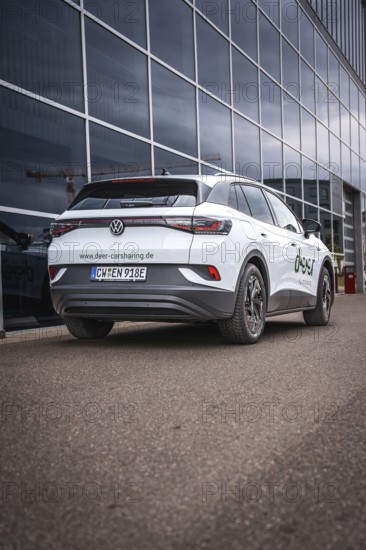 White car in front of glass façade on paved road, VW ID4 electric car, Deer Carsharing, Calw, Germany