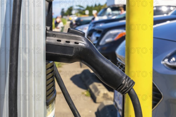 Electric vehicle charger hanging on a charging station, waiting to be plugged into a car, with cars parked in the background in a parking lot