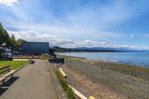 Calm ocean waters lapping a pebbled shore at qualicum beach on vancouver island, british columbia, with benches, buildings, and distant mountains under a bright blue sky