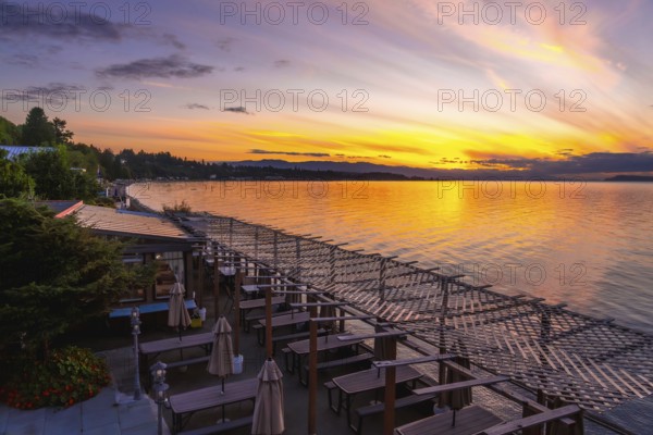 Golden sunset illuminating calm bay waters while reflecting vibrant evening sky colors over a restaurant seating area with a wooden pergola at qualicum beach, vancouver island