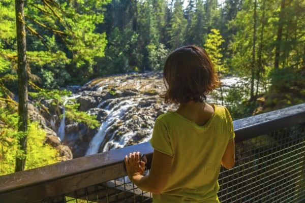 Female tourist standing on a viewpoint, admiring englishman river falls cascading through lush green forest in vancouver island, british columbia, canada, during a sunny summer day