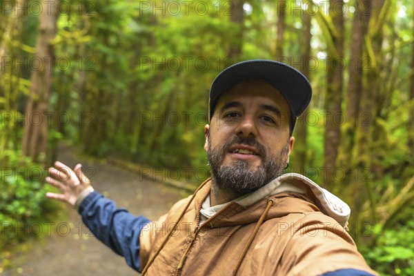 Tourist wearing casual clothing and a baseball cap is gesturing to the rainforest trail while taking a selfie in ucluelet on vancouver island, british columbia, canada