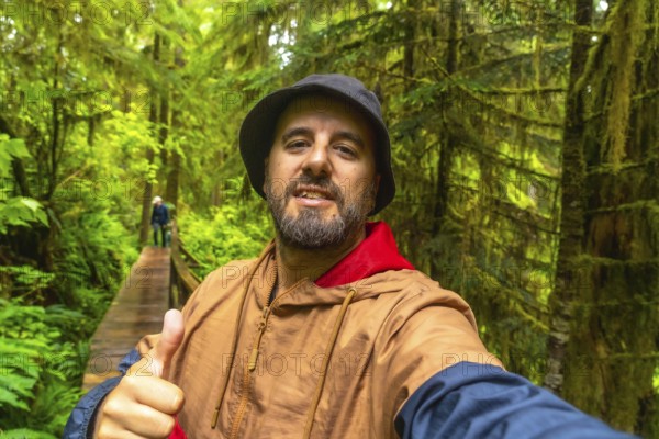 Happy tourist gesturing thumbs up while taking a selfie on a wooden walkway through a lush rainforest in ucluelet, vancouver island, british columbia, enjoying a scenic hike in nature
