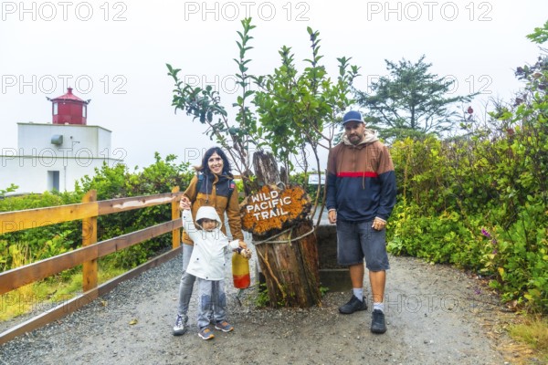 Happy family posing near the wild pacific trail sign with amphitrite point lighthouse in the background, enjoying a cloudy summer day in ucluelet, british columbia, canada