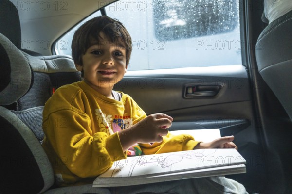 Young child happily drawing in a coloring book while secured in a car seat during a road trip, providing entertainment and creative expression on the journey