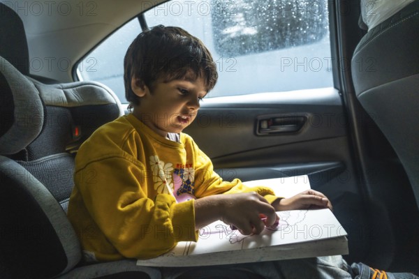 Young child concentrating on creating colorful artwork with crayons while secured in a car seat, enjoying a pastime during a road trip, with a rainy window in the background