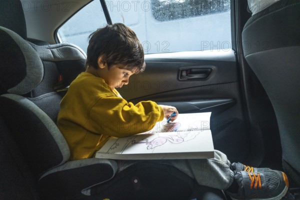 Young child is sitting in a car seat, happily drawing with crayons in a coloring book during a road trip, providing a quiet and engaging activity for travel