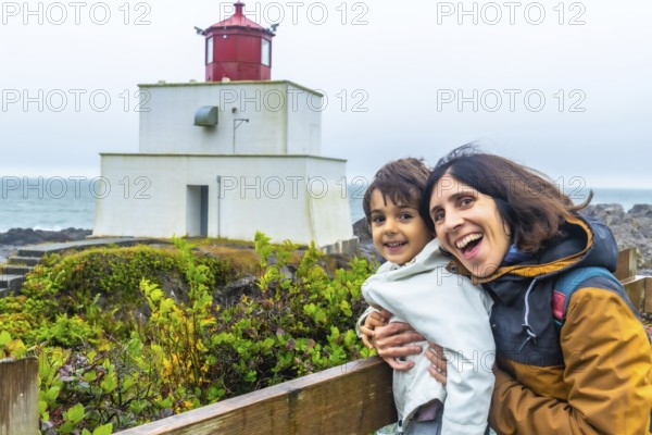 Happy mother and child hugging and smiling with the amphitrite point lighthouse in ucluelet, british columbia, canada, in the background, enjoying a cloudy day by the pacific ocean