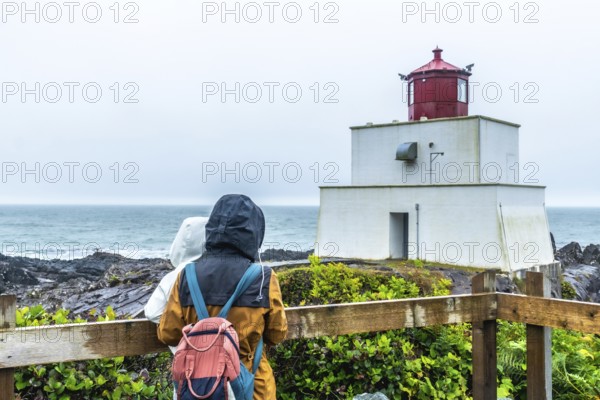 Two tourists in raincoats and backpacks enjoying the misty view of amphitrite point lighthouse, with the ocean and rocky coastline of ucluelet, british columbia, in the background