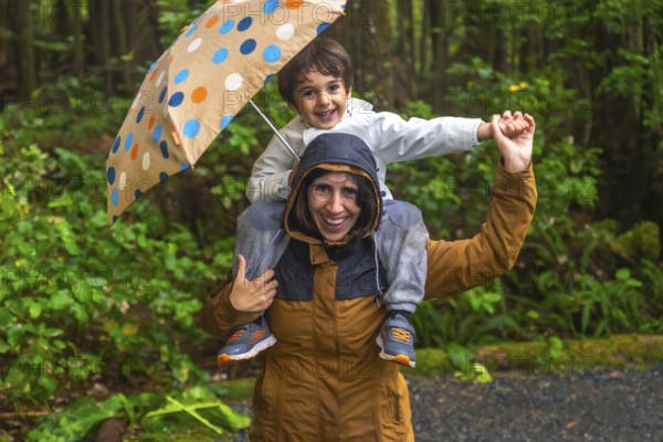 Mother carries her smiling son on her shoulders under a colorful umbrella, both wearing raincoats and enjoying a rainy day adventure in a lush green forest