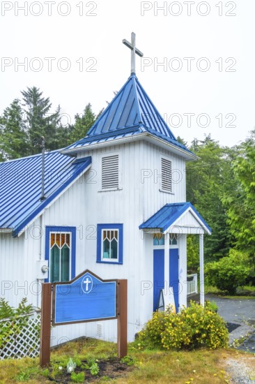 White church building with a blue roof and accents, nestled in ucluelet, british columbia, surrounded by lush greenery and featuring a blank sign in front